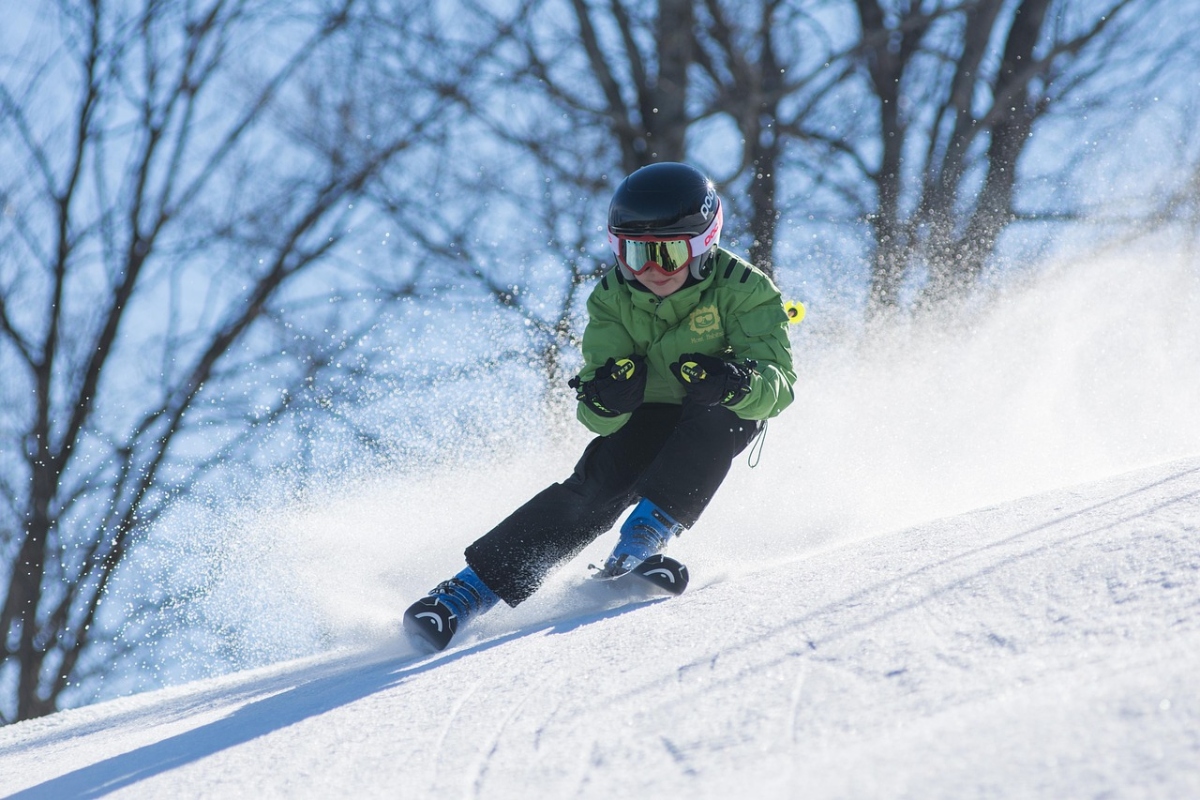 A young skier carves down a snowy slope, wearing a green jacket, black pants, and blue ski boots with a black and pink helmet and reflective goggles. Snow sprays dramatically around the skier as they navigate the turn. Bare trees are visible in the misty background under falling snow.