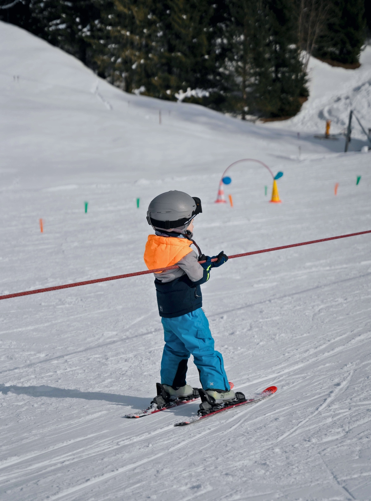 A young child wearing a white helmet, orange and navy jacket, and blue ski pants holds onto a red rope tow while learning to ski on a beginner slope. Colorful training equipment including cones, hoops, and markers are scattered across the groomed snow surface behind the child. Evergreen trees line the gentle slope in the background, with ski trail markers visible on the right side of the image.