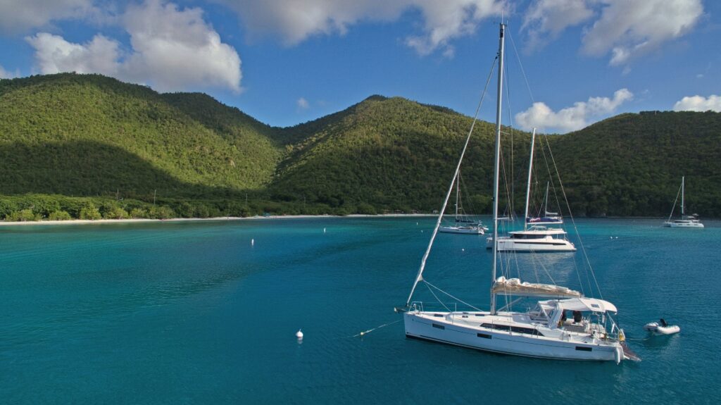 A white sailboat with a tall mast is anchored in the foreground of a sheltered bay filled with brilliant turquoise water, with several other boats and yachts visible at anchor in the distance. The bay is surrounded by densely forested green hills and mountains, with a sandy beach visible along the left shoreline. The scene is captured under a partly cloudy blue sky, creating a picturesque tropical anchorage setting.