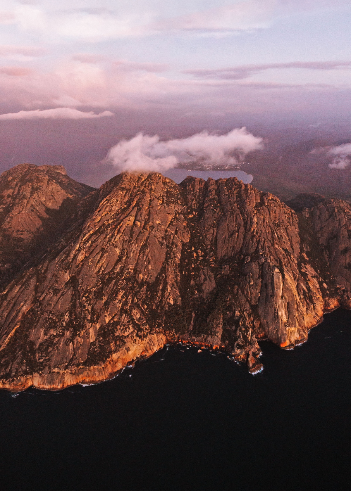 An aerial view shows dramatic, steep granite cliffs and mountain peaks rising directly from dark water, with the rock faces glowing in warm orange and pink tones from golden hour lighting. Low clouds drift around and between the rugged mountain peaks, while a soft pink and purple sky creates a dramatic backdrop. The vertical cliff faces display distinct geological layering and weathering patterns typical of ancient granite formations.