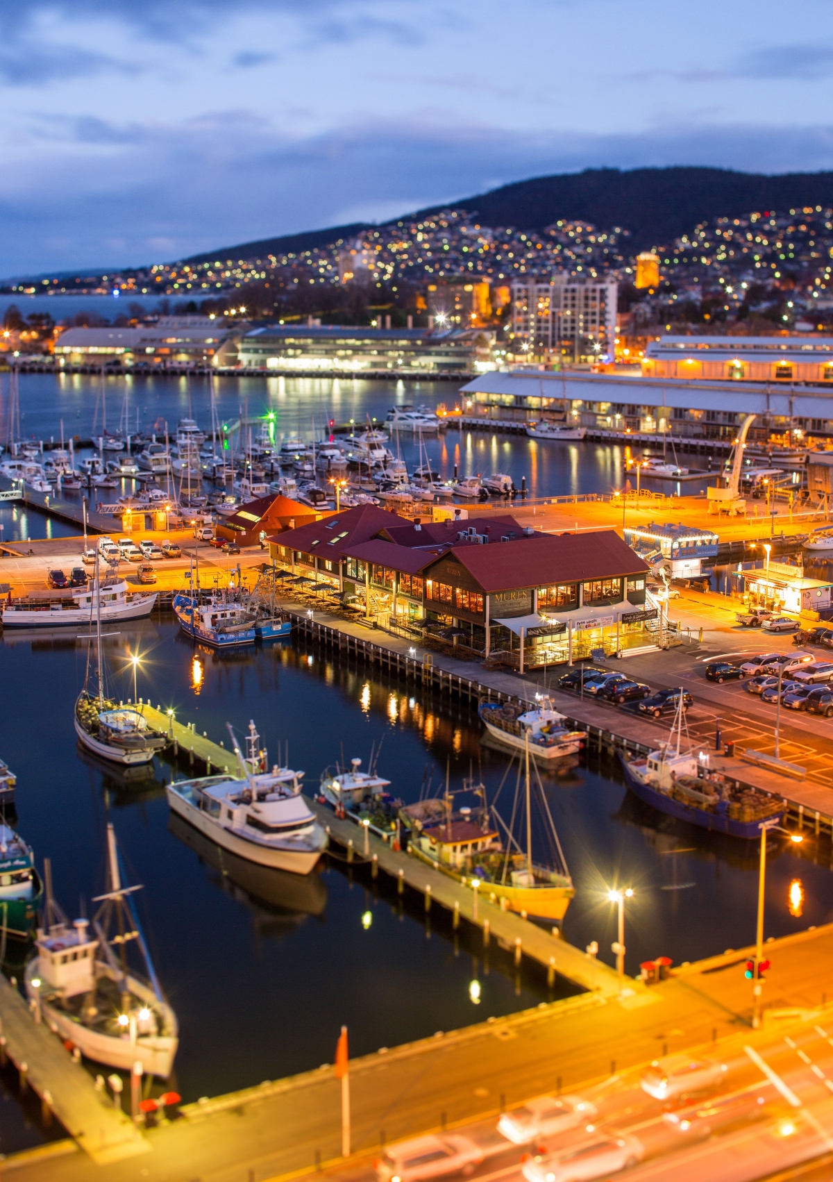 A waterfront harbor scene at dusk shows numerous boats moored at wooden docks and piers, with warm artificial lighting illuminating the marina area. The harbor features waterfront buildings including what appears to be restaurants or commercial structures built over the water, with their lights reflecting on the calm water surface. In the background, a hillside dotted with lights rises behind the urban waterfront, creating a scenic evening cityscape under a cloudy sky.