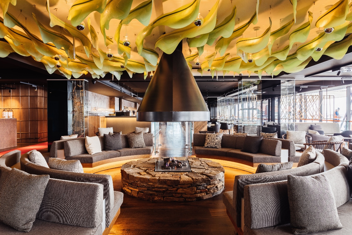 A modern interior lounge features a striking ceiling installation of flowing yellow and green fabric panels suspended overhead. The space centers around a circular stone fireplace with a large conical metal hood, surrounded by grey upholstered sofas and chairs arranged in a conversational circle. The room has an open, contemporary design with warm lighting, exposed ceiling elements, and large windows visible in the background.