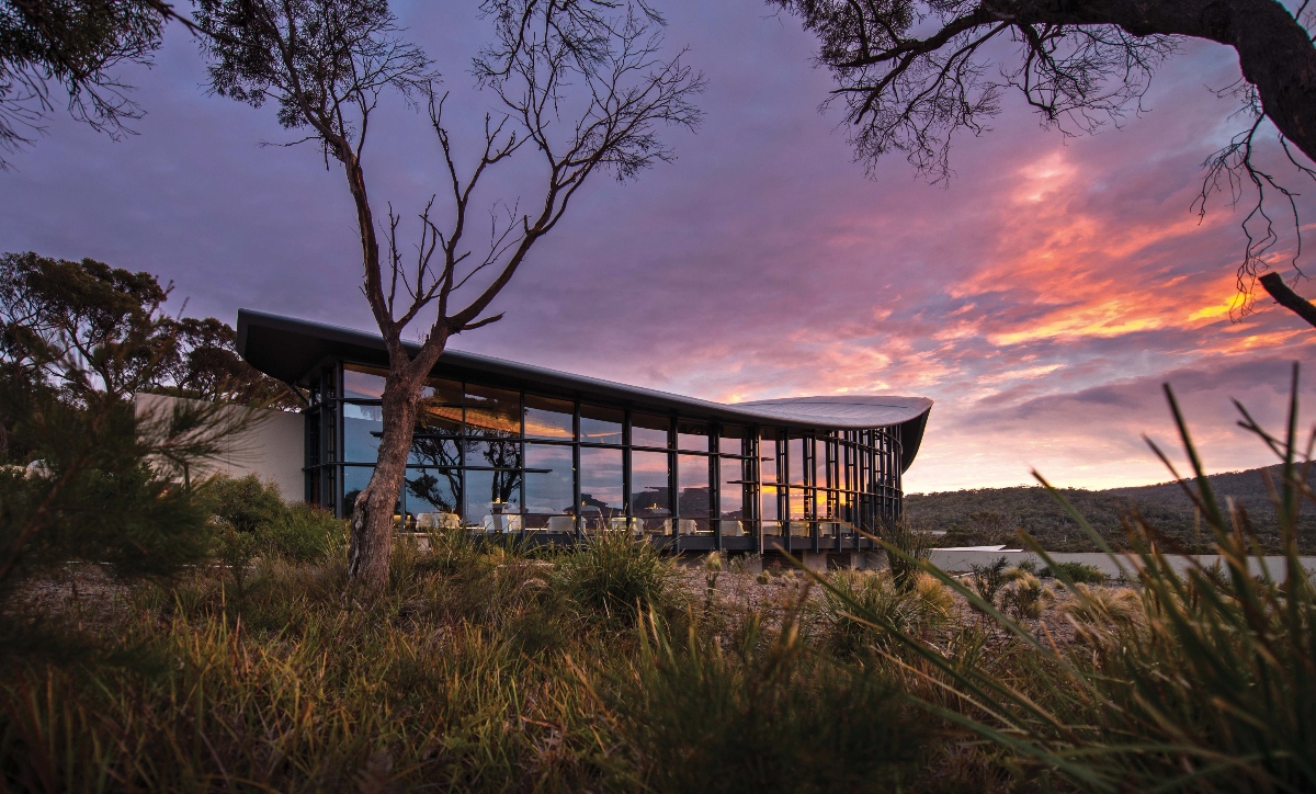 A modern architectural building with extensive floor-to-ceiling glass windows sits nestled among native trees and vegetation in a natural landscape setting. The contemporary structure features a distinctive angular roofline and appears to be positioned on elevated terrain overlooking rolling hills. The scene is captured during golden hour with dramatic purple, pink, and orange clouds filling the sky, while native grasses and a prominent tree frame the composition in the foreground.