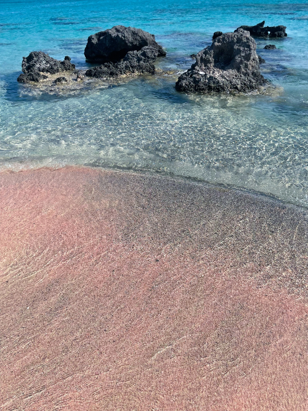 A beach with distinctive pink-tinted sand shows wave patterns and ripples along the shoreline, where clear shallow water meets the shore. Several dark, rough-textured volcanic rocks protrude from the turquoise water, creating contrast against the translucent sea and unusual colored sand.
