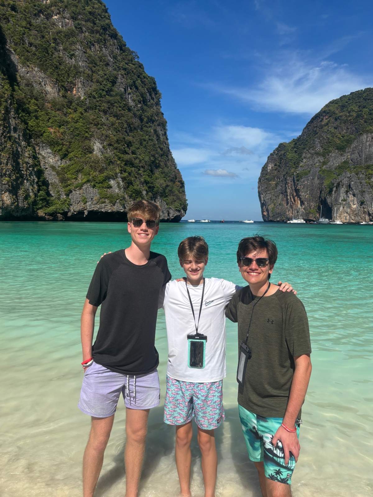 Three young people stand together in shallow turquoise water with their arms around each other's shoulders, wearing casual beachwear and sunglasses. Towering limestone cliffs covered in lush green vegetation frame both sides of the bay behind them, with several boats anchored in the water beneath a blue sky with scattered clouds.