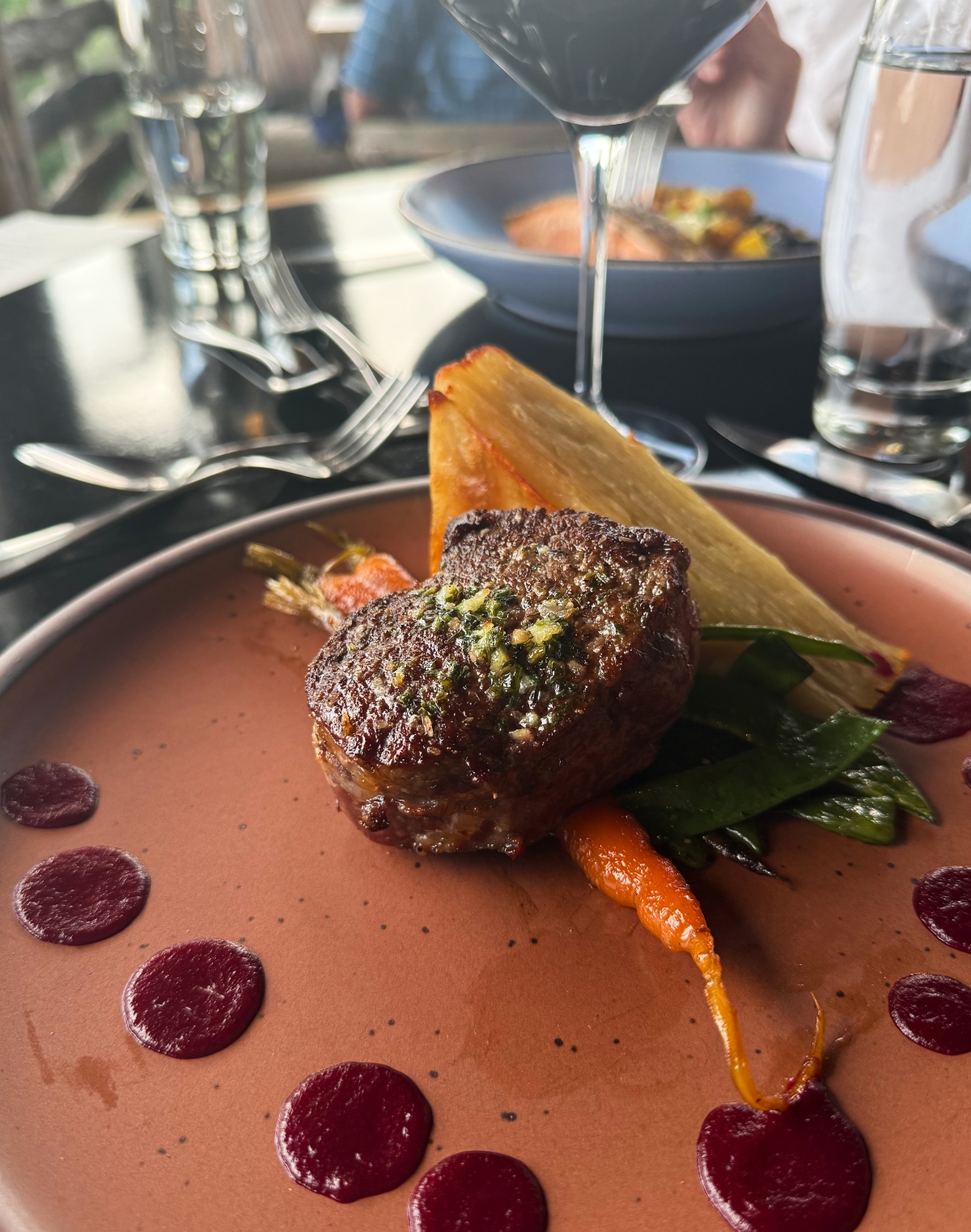 This image shows an elegantly plated restaurant dish featuring a seared piece of meat topped with green herbs, accompanied by colorful vegetables including orange carrots and green beans or snap peas. The plate has a terracotta-colored sauce with dark purple dots arranged around it, and includes a golden triangular potato cake. In the background, table settings with silverware and glassware are visible, and someone can be seen pouring sauce from a small vessel.