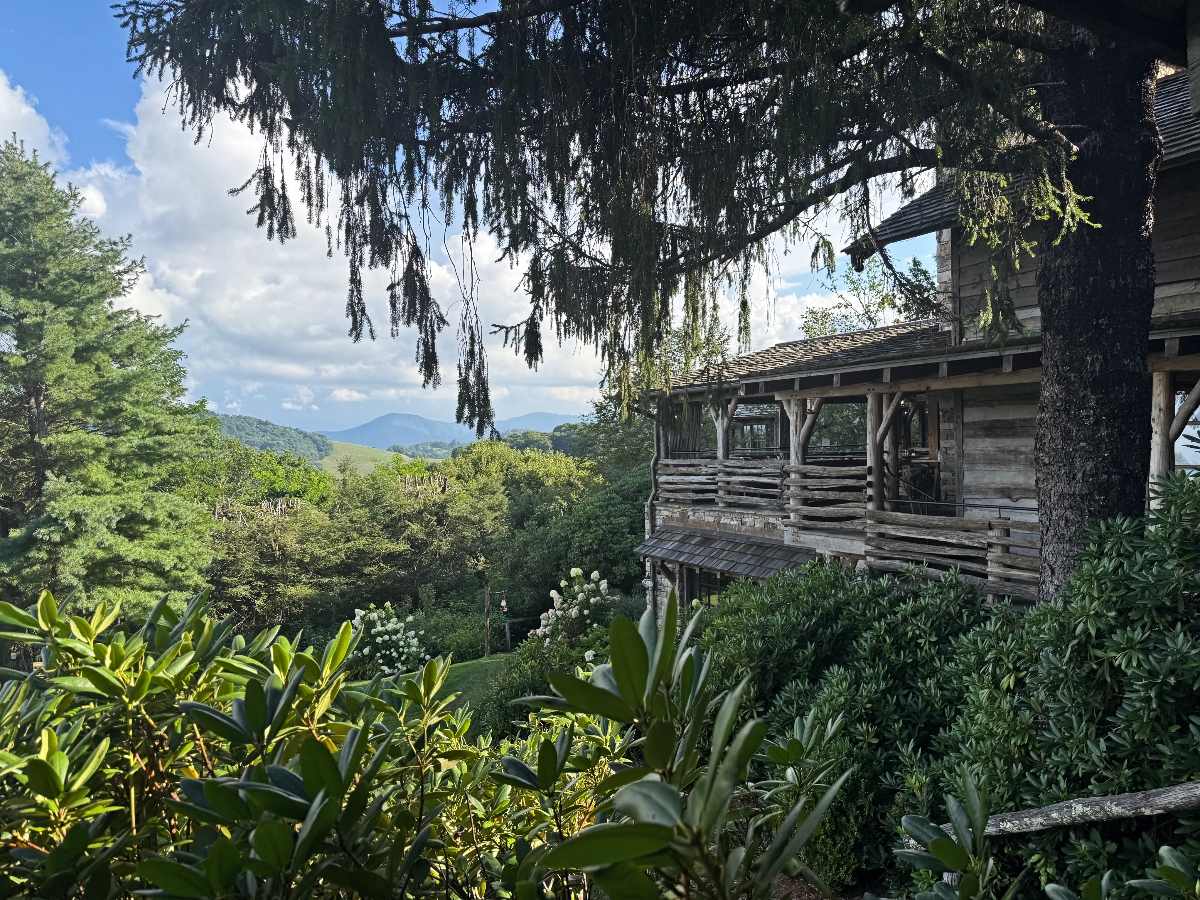 This image shows a rustic wooden cabin or lodge with a covered porch featuring wooden railings and support posts, situated on elevated terrain. The structure is surrounded by lush green vegetation and tropical plants in the foreground, with a large tree with drooping branches prominently featured. In the background, rolling hills and mountains are visible under a partly cloudy blue sky.