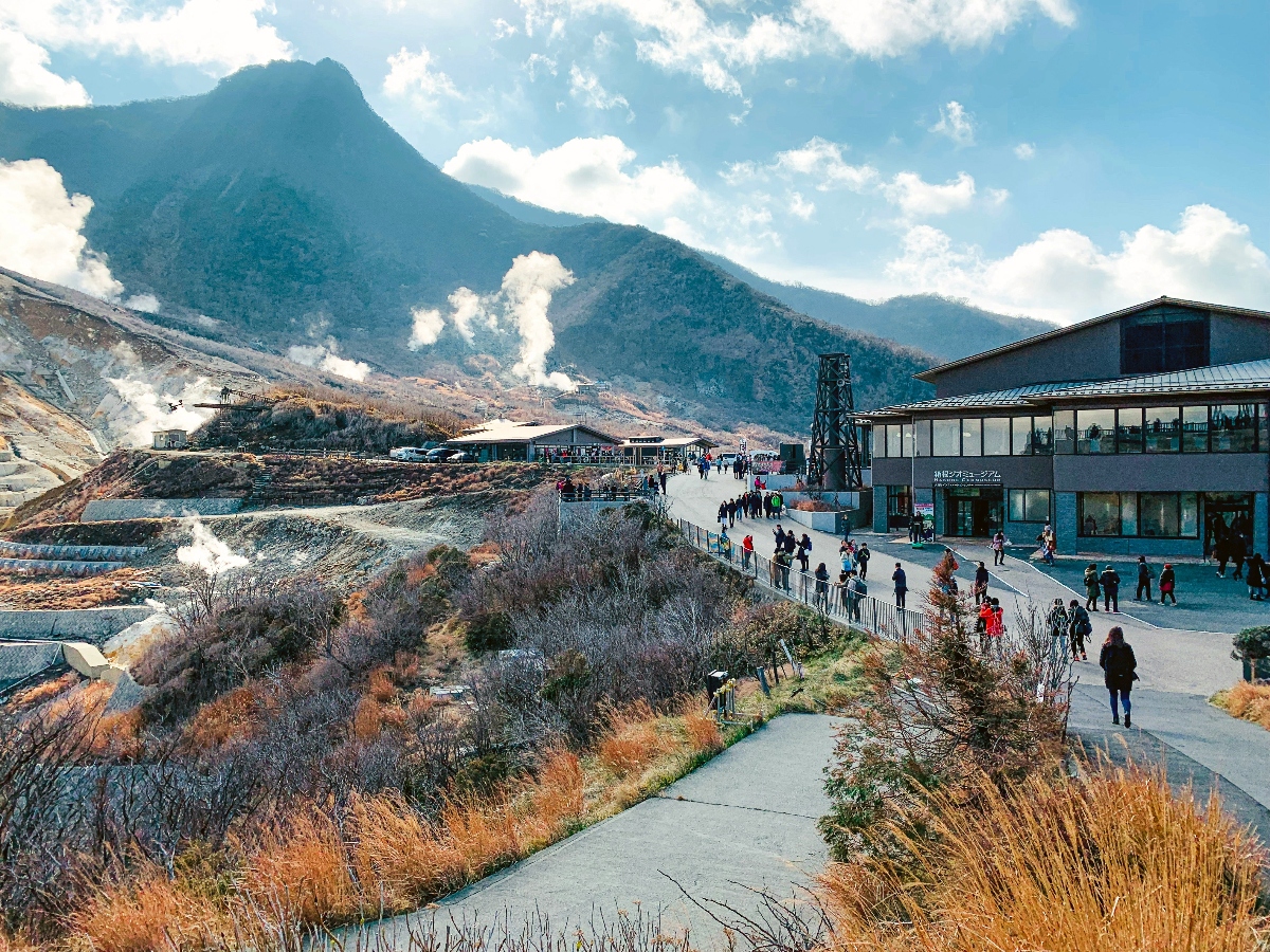 A geothermal area with steam vents rising from barren, rocky terrain in the foreground, set against a backdrop of forested mountains under a partly cloudy sky. A modern two-story building with large windows stands on the right side of a paved walkway, where numerous visitors are walking and gathering. The landscape features dried grasses and sparse vegetation along the pathways leading through the volcanic terrain.
