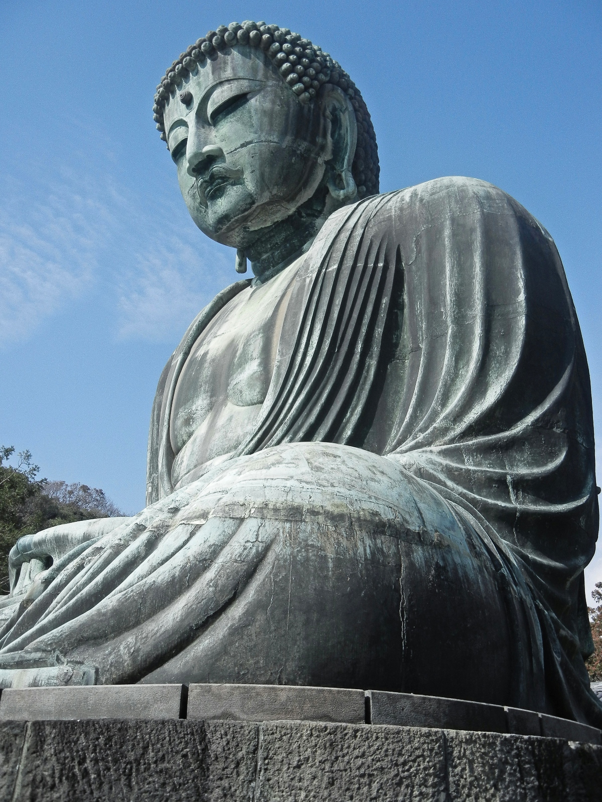 A large bronze Buddha statue photographed from below, showing weathered greenish patina on the metal surface. The statue depicts Buddha in a seated meditation pose wearing flowing robes with detailed drapery folds, with hands resting in the lap and a serene expression with downcast eyes. The monument sits on a stone platform against a clear blue sky, with trees visible in the background.