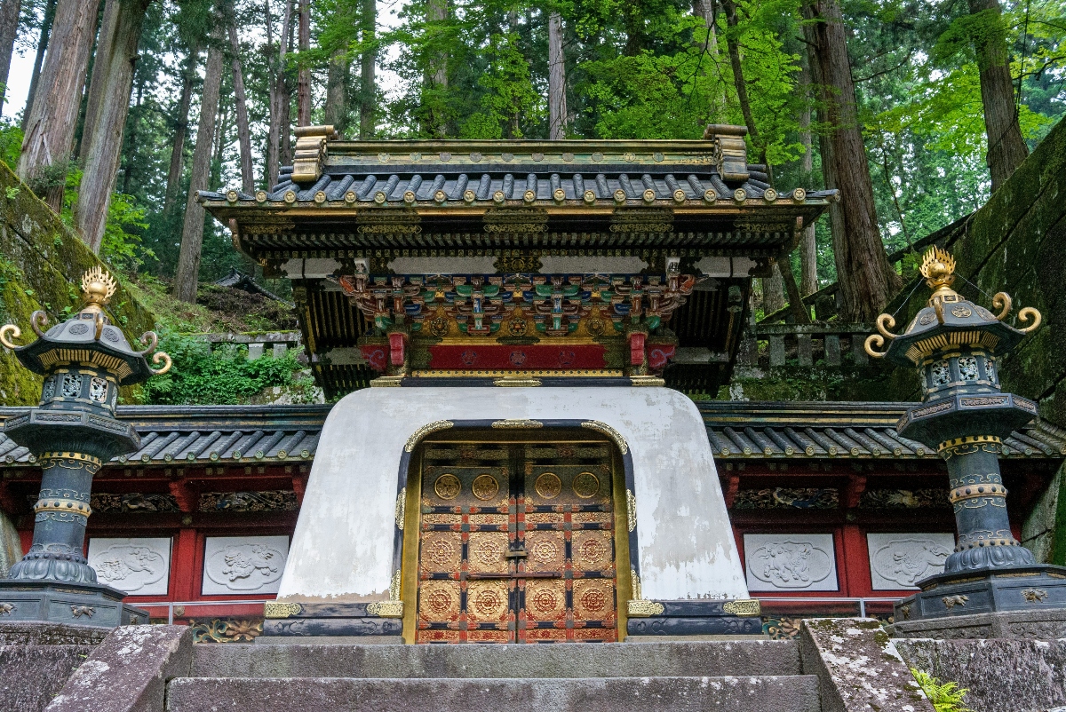 A traditional Japanese temple gate featuring ornate gilded double doors with intricate decorative patterns, framed by a distinctive white curved archway. The structure displays elaborate multi-tiered roofing with dark tiles and colorful carved woodwork in red, black, and gold, flanked by two ornamental bronze lanterns on decorative pedestals. The temple entrance is set within a dense forest of tall trees with vibrant green foliage, accessed by stone steps at the base.