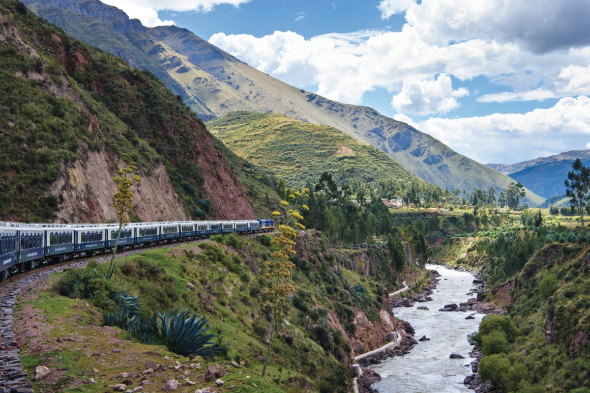 A passenger train with blue and white cars travels along tracks on a steep mountainside, curving through a dramatic Andean landscape with verdant green slopes and exposed reddish-brown rock faces. Below the train, a river flows through a narrow valley flanked by terraced hillsides and scattered vegetation, while towering mountains rise in the background beneath a partly cloudy sky.