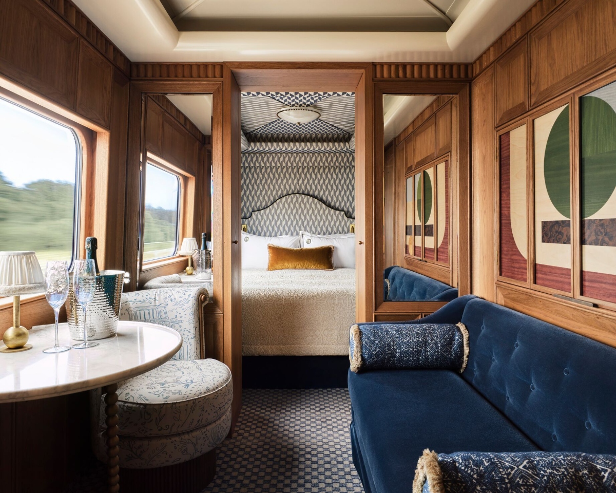 A luxury train cabin features rich wood paneling throughout, with a sleeping alcove containing a bed with geometric patterned headboard and white linens, flanked by large windows showing passing landscape. The cabin includes a small round table set with champagne and glassware near the window, a blue velvet tufted sofa with decorative pillows, and ornate wooden cabinet doors with Art Deco-style inlays on the right wall.