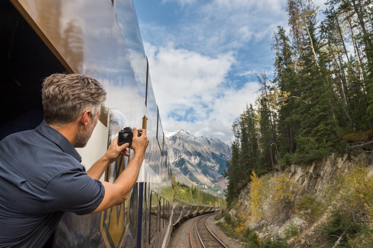 A man in a dark blue shirt leans out of an open train window holding a camera, photographing the scenery as the train curves along tracks through a mountainous landscape. Snow-capped peaks rise in the distance beyond dense evergreen forests and rocky cliff faces that line the railway route under a partly cloudy sky.