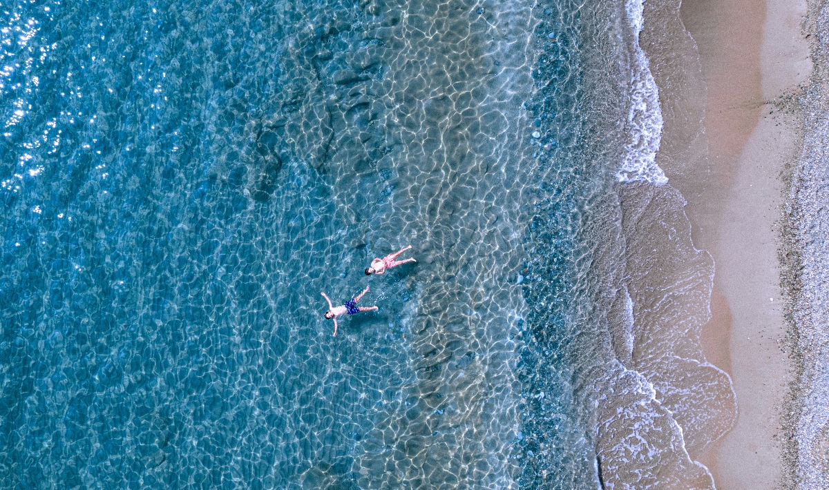 An aerial view shows two people floating in crystal-clear turquoise water near a sandy shoreline, with one person wearing pink swimwear and another in dark swimwear. The transparent water reveals light patterns reflecting on the sandy ocean floor, while white foam from gentle waves marks the boundary between the deep blue sea and the pale sandy beach.