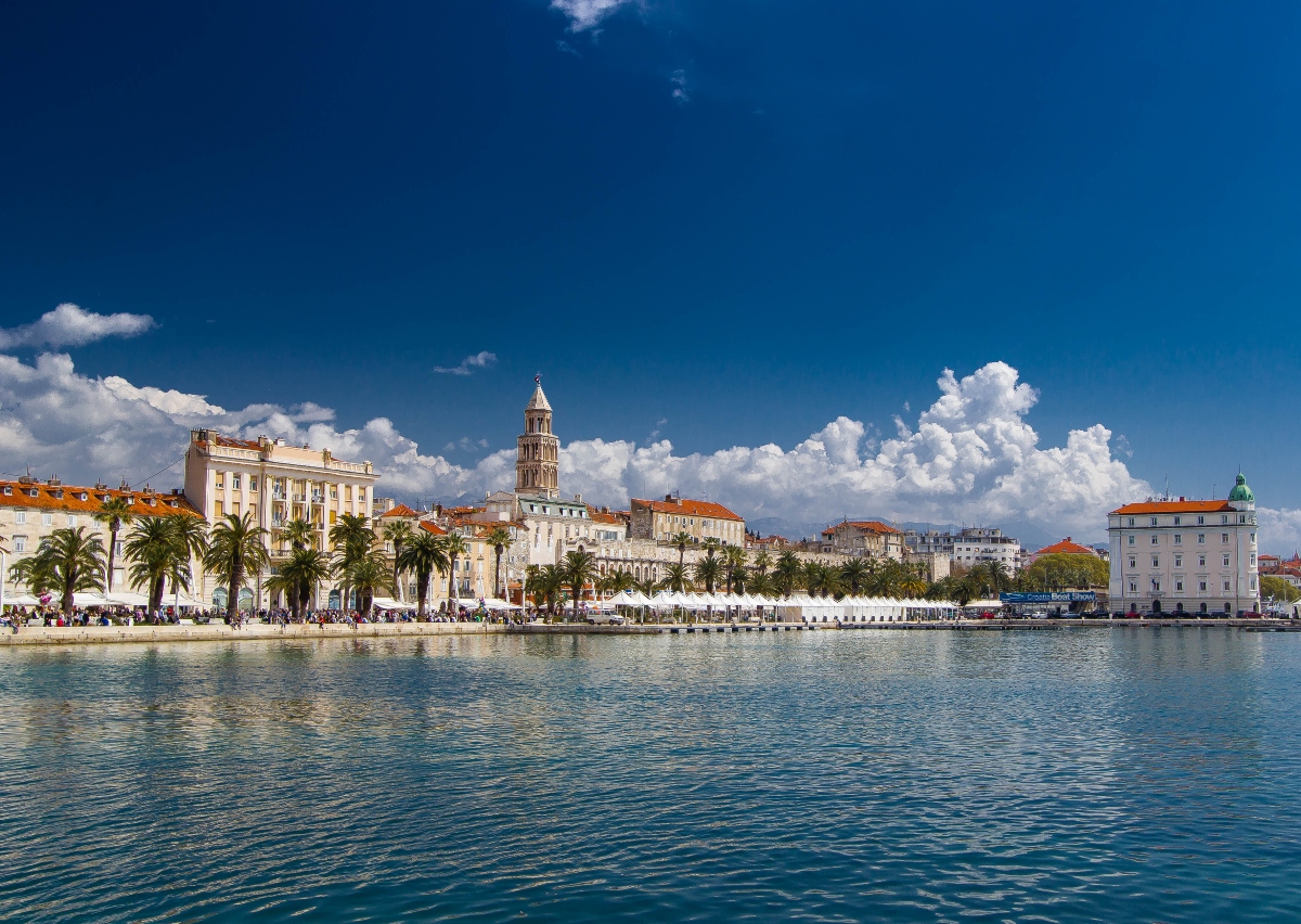 A waterfront view shows calm blue water in the foreground with a stone promenade lined with palm trees along the shore, where numerous people can be seen walking. Behind the promenade stands a row of historic Mediterranean-style buildings with cream and white facades and red tile roofs, dominated by a tall stone bell tower rising prominently against a blue sky with white clouds.