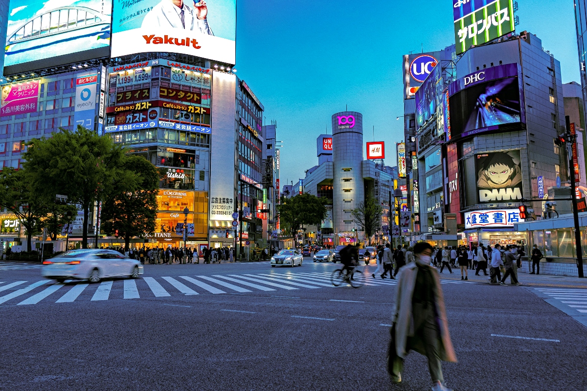 A busy pedestrian intersection with wide zebra crossings where numerous people walk across the street during evening hours, with motion blur capturing the movement of pedestrians and vehicles. The scene is surrounded by tall commercial buildings displaying large illuminated digital billboards and signage, including recognizable brands, creating a vibrant urban landscape typical of Tokyo's Shibuya district, one of the best vacation ideas for teens.